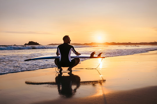 Surfer with surfboard sits on ocean surf line at sunset time