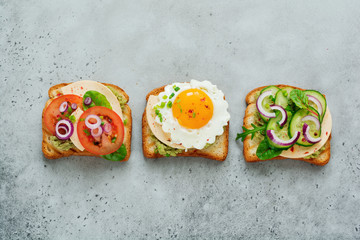 Three different toast sandwiches with guacamole sauce, fried egg, fresh cucumber, cheese and tomato with chard, arugula, red onion on gray concrete background. Selective focus.