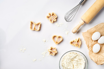 Bowl with wheat flour, rolling pin, whisk, eggs, cookie cutters. Top view on a white table with a...