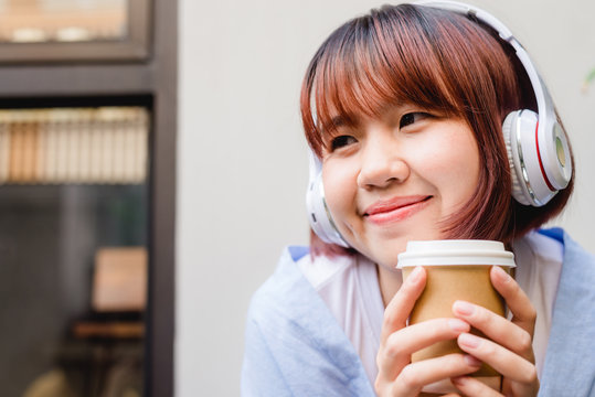 Happy young Asian woman listening to music in a coffee shop holding a cup of coffee in her hand. Young Woman listening to music with headphone while relaxing in the garden. Coffee relax concept.
