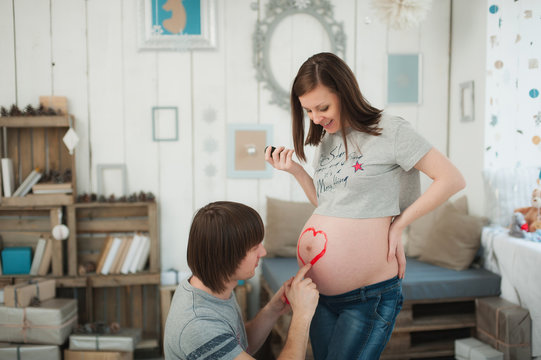 Pregnant woman with big belly. man paints heart with paints on tummy. Couple with kid clothes. Wife, husband. Pregnancy, parenthood, family, motherhood, parents, children, people, expectation concept.