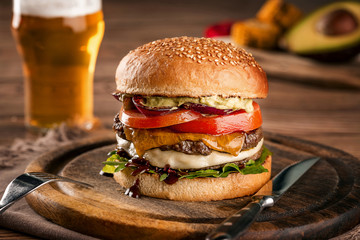 Cheeseburger, made from rye bun with tomato slice and melted cheese on a roasted beef, and lettuce leaf on a wooden table, with a glass of beer