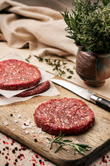 On a wooden cutting board on kraft paper there are raw beef burgers for burgers, spices, rosemary, a pepper mill, a cooking process. Top view