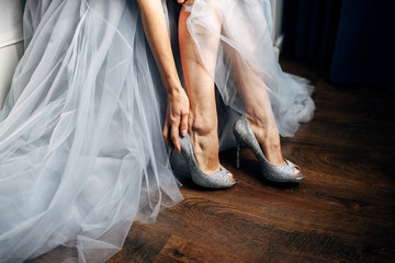 Bride puts on her silver wedding shoes, grey wedding dress on background