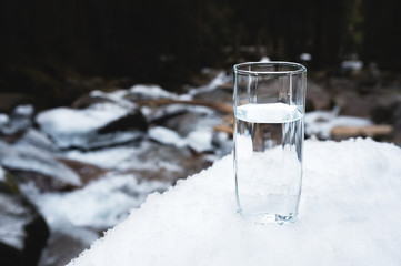 A transparent glass glass with drinking mountain water stands in the snow against a background of a clean frost mountain river in winter.