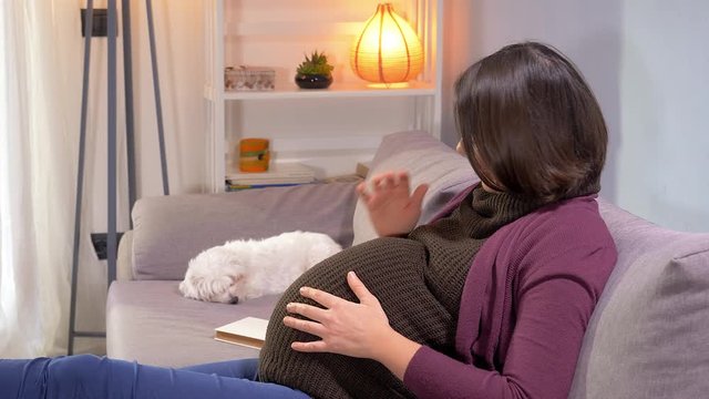 Beautiful pregnant woman feeling kick of baby sitting on sofa at home happy