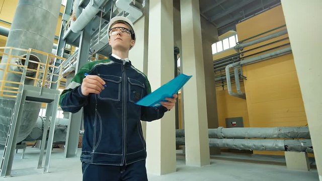 Engineer in hardhat walking in factory and writing down on clipboard