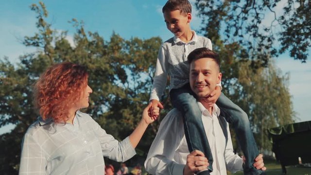 Cheerful young family walking down the park, father carrying his son on shoulders, a boy holds his mother’s hand, they have a pleasant conversation. Positive emotions, family, happy childhood