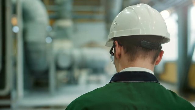 Backview of engineer in hardhat walking in factory and writing down on clipboard