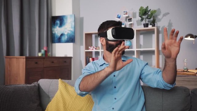 young man wearing virtual reality glasses waving his hands sit on the sofa at home feel happy shock innovation looking modern entertainment gadget new tech visual beard game hipster slow motion