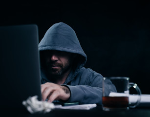 male hacker in a sweatshirt with a hood sitting at a table and looking at the laptop