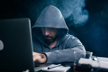 a man hacker in a sweatshirt with a hood is sitting at a table and something is typing in a laptop