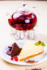 Cheesecake decorated with chocolate pieces, cranberry and mint leaves on white plate and glass teapot