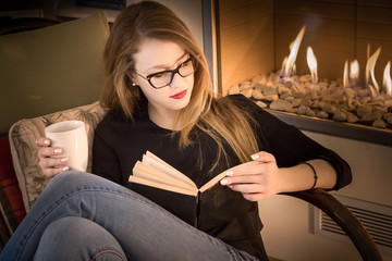 Portrait of a young blonde woman wearing eyeglasses, reading a book at the fireplace, indoors.