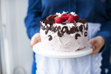 A woman is holding a beautiful cake with pink cream and fresh raspberry berries, decorated with chocolate. Calorie food. Free space for text or postcard.
