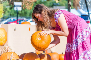 Young woman in pink dress picking up holding heavy large pumpkins from group stack in outdoor market store for holidays in country countryside