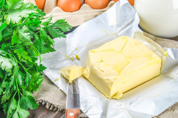 A bar of butter is cut into pieces on a wooden board with a knife, surrounded by milk, eggs and parsley on a brown table. Ingredients for cooking.