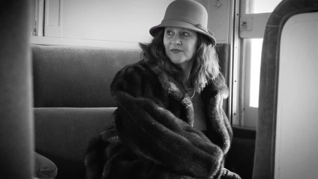 A lovely passenger train traveler in stylish 1930s or 1940s period attire sitting in a seat of a vintage railcar looks at her ticket.