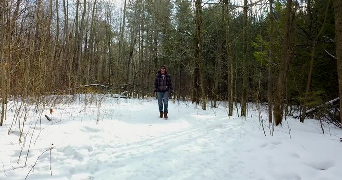 Drone shot of a young man walking in a snowy forest, his dog hangs back on the path behind him.  4K.