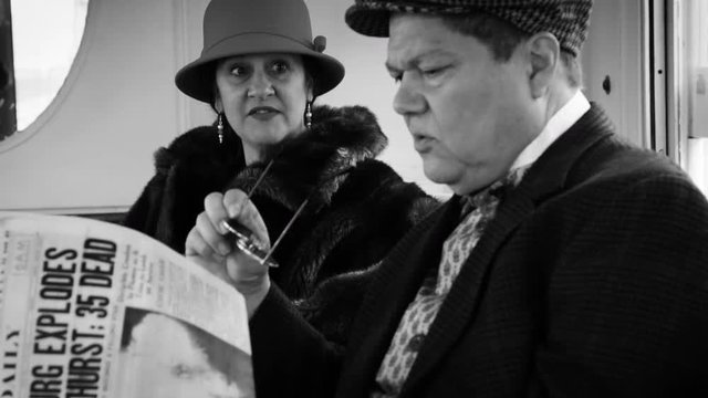 A man and woman traveling on a passenger train dressed in stylish 1930s or 1940s period attire sitting in the lounge car of a vintage railcar reading and discussing a news paper article.
