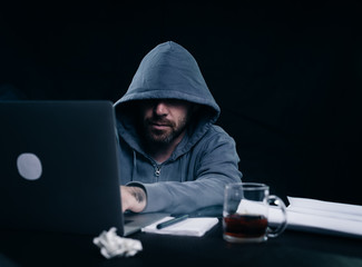 a man hacker in a sweatshirt with a hood is sitting at a table and something is typing in a laptop