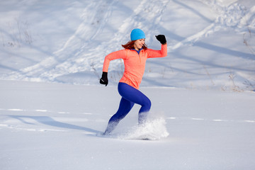 Fototapeta na wymiar A young woman in a bright blue hat, an orange sweatshirt and elk skirts runs through the winter snow on a bright winter day, a side view