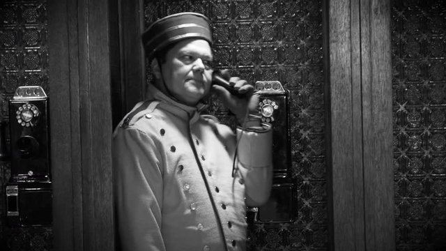 An old timey type bellhop or porter in a vintage phone booth impatiently listening to someone on the other line.