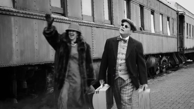 Period piece 1930s_1940s passenger train travelers in vintage attire carrying luggage walking next to railcars and upon arrival greeting someone in the distance.