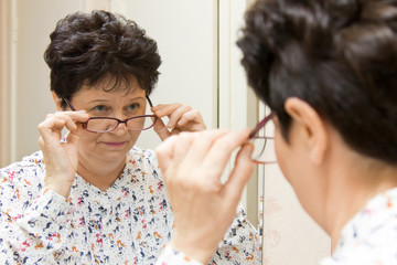 Senior woman trying on new eyeglasses and looking over the glasses at herself in the mirror 