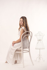 Young girl with sheer skin and Nude makeup sits on a white vintage chair on a grey isolate