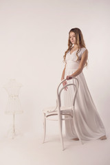 Fototapeta premium A young girl in a long white dress and Nude makeup standing in front of a vintage white chair on the gray isolate