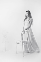 A young girl in a long white dress and Nude makeup standing in front of a vintage white chair on the gray isolate black and white image