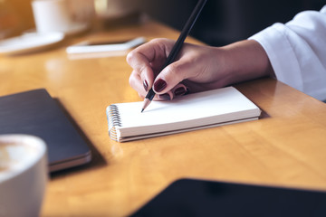 Closeup image of woman's hand writing on a blank notebook with laptop , tablet and coffee cup on wooden table background