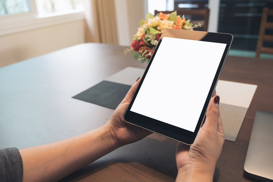 Mockup image of hands holding black tablet pc with blank desktop white screen on wooden table in the house