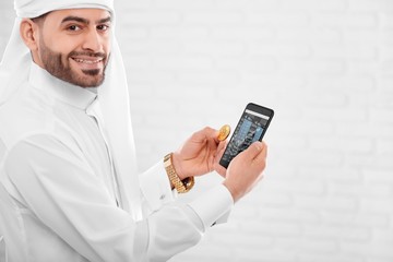Young Muslim man keeps golden bitcoin and mobile phone in another hand. He stands on the white studio background. A businessman looks happy and engaged.
