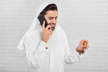 A Muslim businessman wearing traditional white costume, looks on golden bitcoin in his hand. In another hand he keeps his black mobile phone. He wears golden watch and cufflinks on his shirt.