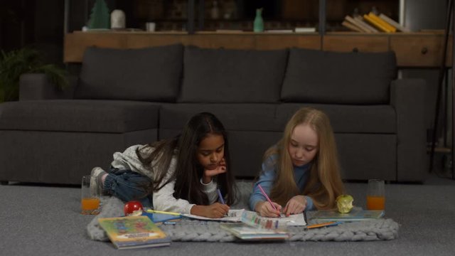 Beautiful multiracial little girls painting coloring pages for kids with colorful markers while sitting on the floor. Happy school girls making drawings and paintings together in domestic room.