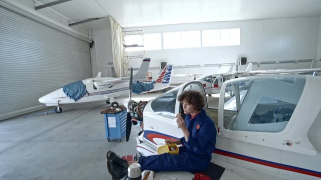 Tilt down of female aircraft mechanic sitting on wing of light jet airplane in hangar, eating vegetable salad from container and reading manual