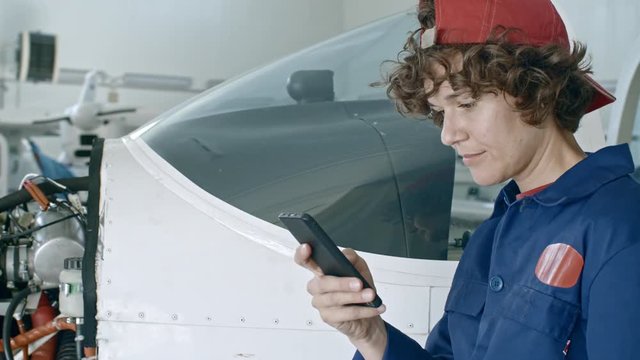 Female aircraft mechanic standing near jet airplane in hangar, texting on smartphone and then looking to the side