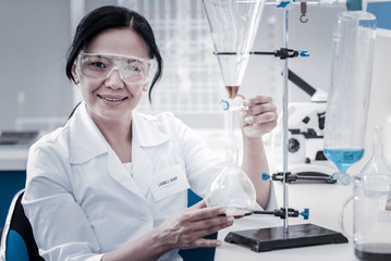 Fototapeta premium Loving my job. Selective focus on a beautiful mature lady wearing a labcoat and safety glasses looking into the camera with a cheerful smile on her face while touching glassware fixed on a stand.