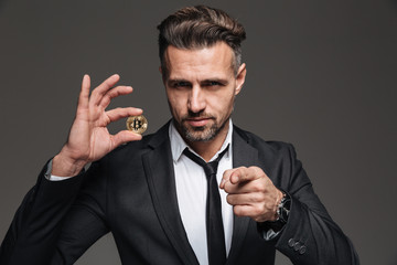 Image of successful male entrepreneur in business suit and tie holding crypto currency and pointing finger on camera, isolated over dark gray wall