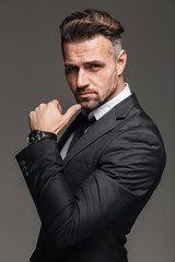 Portrait of rich brunette man 30s in black suit posing on camera with stylish watch on wrist, isolated over graphite background