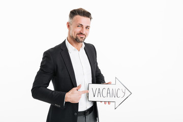 Photo of smiling successful man wearing black jacket gesturing on speech arrow pointer with word vacancy in hand, isolated over white background