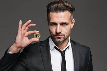 Photo of successful mature man with brown hair in suit and tie demonstrating golden bitcoin on camera, isolated over dark gray background