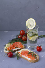 sandwich with red fish trout a piece of lemon and delicate cheese a glass of water with lemon mint and ice juicy red cherry tomatoes on a stone table