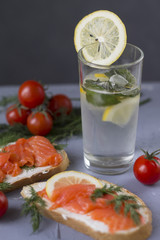 sandwich with red fish trout a piece of lemon and delicate cheese a glass of water with lemon mint and ice juicy red cherry tomatoes on a stone table
