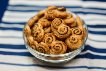 A horizontal lifestyle image of a glass bowl filled with homemade cookies, made of puff pastry, with sugar and cinnamon. On a surface, covered with blue and white stripy towel. Selective focus.