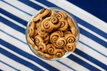 A horizontal lifestyle image of a glass bowl filled with homemade cookies, made of puff pastry, with sugar and cinnamon. On a surface, covered with blue and white stripy towel. Selective focus.