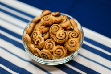 A horizontal lifestyle image of a glass bowl filled with homemade cookies, made of puff pastry, with sugar and cinnamon. On a surface, covered with blue and white stripy towel. Selective focus.