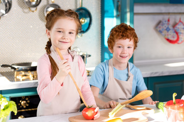 adorable happy children in aprons smiling at camera while cooking together in kitchen
